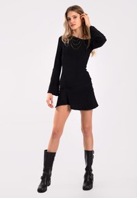 Schwarzes Mini-Kleid mit langen, ausgestellten Ärmeln, gerafft an der Seite und einem leicht asymmetrischen Saum. Getragen mit kniehohen schwarzen Stiefeln.