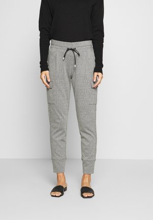 Personne portant un pantalon de jogging gris avec des poches latérales, un haut à manches longues noir et des sandales à talons ouvertes noires, debout sur un sol simple.