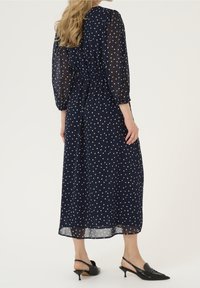Robe maxi bleu marine à pois blancs, manches longues transparentes, taille smockée et silhouette fluide. Portée avec des escarpins noirs à bride arrière.
