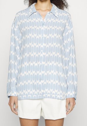 Persona che indossa una camicia azzurra e bianca con fantasia, maniche lunghe e pantaloncini bianchi.