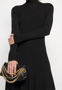 Svart ribbad stickad klänning med långärm och hög hals, med en figurnära design. Accessoarer inkluderar en svart väska med guldkedjedetaljer.