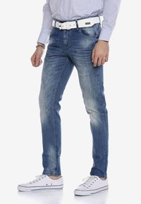 Hellblaue Denim-Jeans, Slim Fit, mit Ausbleichungen. Kombiniert mit weißen Sneakers mit rotem Streifen. Ein weißer Gürtel ergänzt das Outfit.