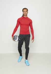 Rotes langärmliges Sportshirt, schwarze Sporthose mit weißen Streifen, blaue Sportschuhe, hält einen schwarz-weißen Fußball.