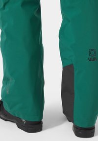 Grüne wasserdichte Hosen mit einem schwarzen unteren Bereich, geradem Schnitt und Logodetails am Bein, kombiniert mit robustem schwarzen Schuhwerk.