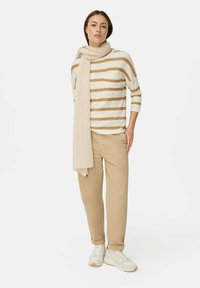 Gestreiftes, beige und cremefarbenes Strickoberteil mit einer Fronttasche, kombiniert mit hellbraunen Hosen und einem passenden Schal. Weiße Sneaker runden das Outfit ab.