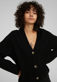 Schwarze gerippte Strickjacke mit tiefem V-Ausschnitt, übergroßen Ärmeln und fünf goldfarbenen Knöpfen auf der Vorderseite. Strukturierter Stoff mit entspannter Passform.