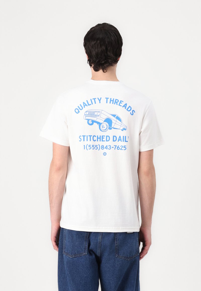 White cotton t-shirt featuring blue graphic print on the back with text "QUALITY THREADS," a car illustration, and contact number.