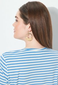 Zlaté a tkané náušnice s kruhovým a vějířovitým designem, nošené s modro-bílým pruhovaným tričkem, profilový pohled na model.