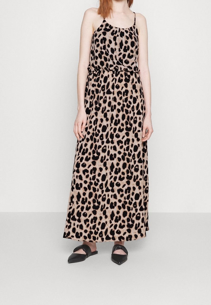 Femme portant une robe maxi sans manches beige et noire à imprimé léopard, avec des fines bretelles et des chaussures plates noires pointues.
