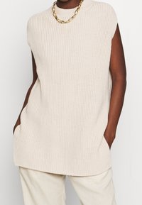 Pletený bezrukávový top ve světle béžové barvě s žebrovanou texturou, bočními kapsami a vysokým límcem. Nošen přes světlé kalhoty.