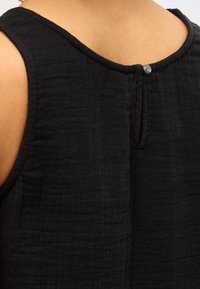 Κοντινή λήψη στο επάνω μέρος της πλάτης ενός ατόμου που φοράει μαύρο υφασμάτινο αμάνικο τοπ με μικρό κουμπί και κούμπωμα στο ντεκολτέ.