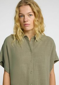 Olive green button-up shirt made of lightweight fabric, featuring a textured surface and short sleeves with a relaxed fit.