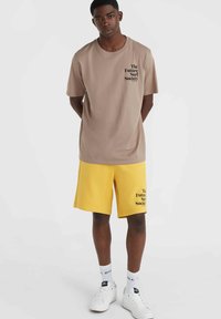 Brown cotton t-shirt with black text, paired with yellow shorts featuring the same branding. White sneakers and patterned socks complete the look.