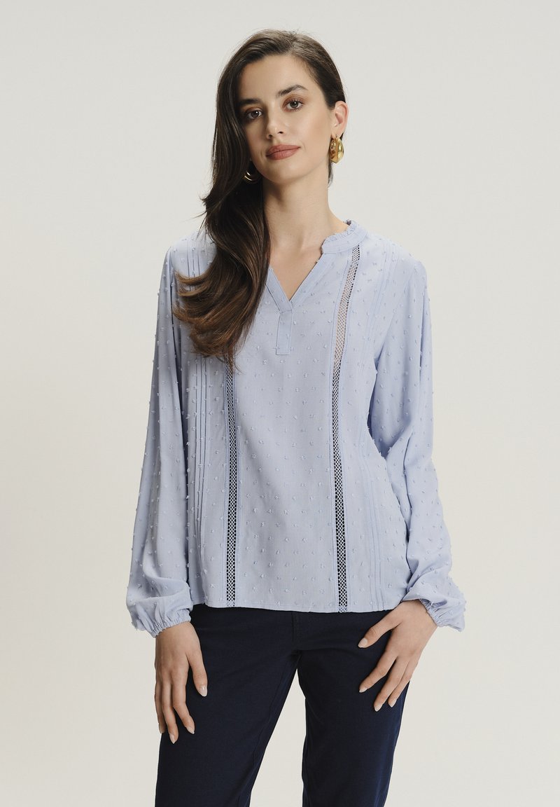 Blusa de manga larga azul claro con escote en V, tela texturizada, patrón de puntos sutil y detalles de encaje a lo largo de las costuras verticales.