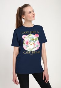 Marinblå t-shirt med en färgglad blommig Care Bear-grafik och gyllene text som säger "CARE LIKE A CARE BEAR 1982." Kort ärm, rund hals.