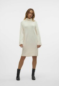Abito a maglia color crema, lungo fino al ginocchio, con colletto alto, maniche a coste e vestibilità comoda. Indossato con stivali neri alla caviglia.