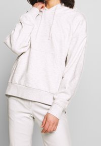 Hellgrauer Kapuzenpullover mit einer Fronttasche, Kordelzugkapuze und gerippten Bündchen. Weiche Stoffstruktur mit lockerem Schnitt. Kombiniert mit glänzenden weißen Hosen.