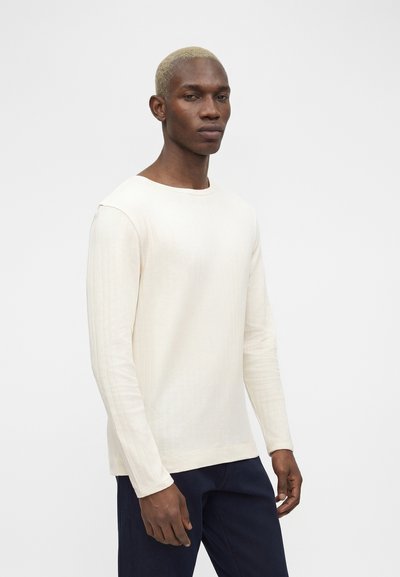 Άνδρας με κοντά ξανθά μαλλιά, που φοράει μια μακρυμάνικη κρεμ μπλούζα και σκούρο παντελόνι, στέκεται μπροστά σε ένα απλό φωτεινό φόντο.