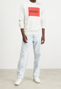 Felpa bianca con logo rettangolare rosso "HUGO" in nero, abbinata a jeans azzurri chiari e sneakers bianche, in piedi su uno sfondo neutro.