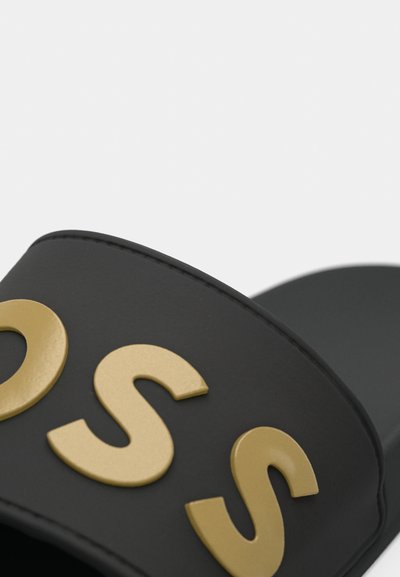 Gros plan d'une sandale noire à semelle plate avec des lettres dorées « OSS » en relief sur la sangle, sur un fond clair.