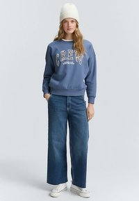 Plava majica s natpisom "CREW" u uzorkovanim slovima, u kombinaciji s trapericama širokih nogavica i bijelim tenisicama. Model nosi bijelu pletenu kapu.