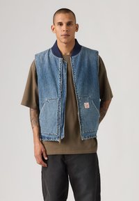 Άνδρας με κοντά μαλλιά, φοράει μπλε τζιν γιλέκο πάνω από καφέ φαρδιά μπλούζα και μαύρο παντελόνι, στέκεται με τα χέρια εν μέρει στις τσέπες.