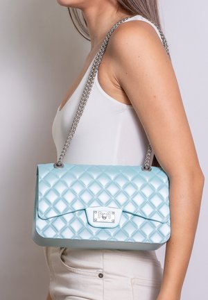 Femme portant un débardeur blanc et un pantalon clair, portant un sac à bandoulière matelassé bleu clair avec une chaîne argentée.