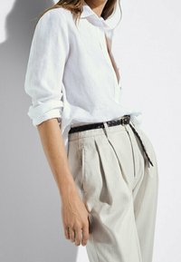 Camicia di lino bianca con maniche arrotolate abbinata a pantaloni beige chiaro a pieghe, fissati con una cintura intrecciata nera. Tessuto morbido.