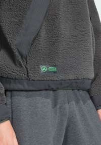 Grå fleecepullover med ribbede detaljer, udstyret med et grønt logo. Tekstureret materiale med rundet kant, parret med matchende grå bukser.