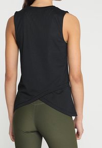 Černý tílko bez rukávů s volnějším střihem, s překříženým zadním dílem a lehkou, prodyšnou texturou. Kombinováno s olivově zelenými legínami.