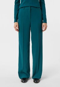 Spodnie o szerokich nogawkach w kolorze morskim z gładkiego materiału, z płaskim przodem i subtelnymi szlufkami na pasek. W połączeniu z czarnymi butami.