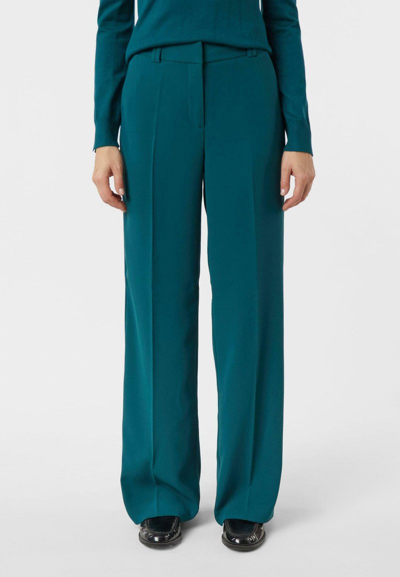 Spodnie o szerokich nogawkach w kolorze morskim z gładkiego materiału, z płaskim przodem i subtelnymi szlufkami na pasek. W połączeniu z czarnymi butami.