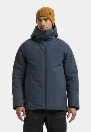 Man wearing a dark blue hooded winter jacket and a mustard yellow knit beanie, standing with hands relaxed at sides.