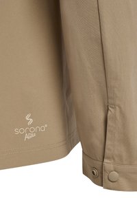Zbliżenie beżowej tkaniny z logo Sorona Agile i częścią mankietu z zatrzaskiem na ubraniu.
