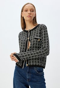 Cardigan à motif noir et blanc avec des accents de boutons dorés, en maille texturée, à encolure ronde et avec des poignets festonnés.