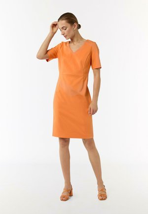 Oranges knielanges Kleid mit kurzen Ärmeln und V-Ausschnitt; es verfügt über einen taillierten Oberteil, subtile Nähte und eine glatte Textur. Kombiniert mit orangefarbenen High Heels.