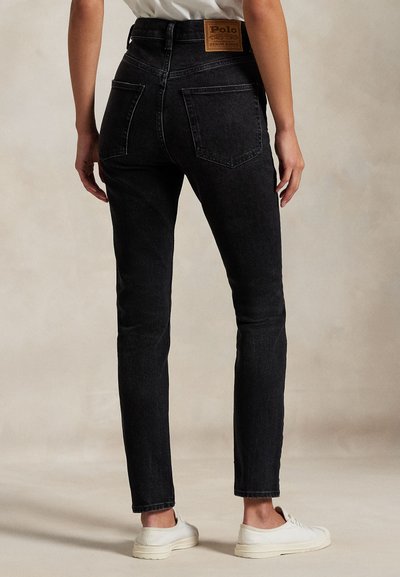Persoon die zwarte high-waisted jeans en witte sneakers draagt, staand met zichtbare rug tegen een eenvoudige neutrale achtergrond.
