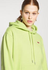 Lys grøn hettegenser laget av myk tekstil med kengurulomme, snor og en liten rød Levi's-logo på venstre bryst.