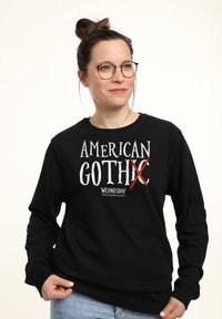 Una donna con occhiali e capelli raccolti in un bun indossa un felpa nera con il testo "American GothX Wednesday", con una X rossa che attraversa parte della parola "Gothic".