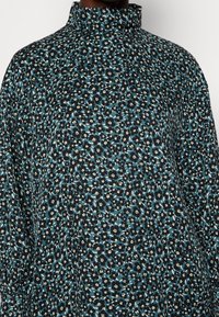 Top à imprimé floral bleu foncé, avec un col haut et une silhouette ample. Le matériau a une texture lisse avec de petites fleurs blanches et vertes.