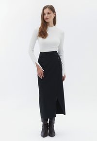 Falda negra envolvente con detalle de botones, combinada con una camiseta de manga larga de canalé blanca ajustada. La modelo lleva botines marrones. Diseño minimalista.