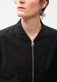 Veste bomber noire avec un col rond, dotée d'une fermeture éclair à l'avant et de deux poches. Fabriquée en tissu texturé avec un design minimaliste.
