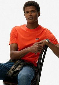 Pomarańczowa bawełniana koszulka z okrągłym dekoltem i małym logo. Model siedzi na krześle, ubrany w niebieskie dżinsy i koszulę w kratę związaną w talii.