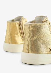 Złote metaliczne buty typu high-top z bocznym zamkiem błyskawicznym, białą gumową podeszwą i fakturowaną powierzchnią. Posiadają kontrastowe szwy i akcenty w postaci oczek.