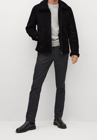 Sort imitation pelsjakke, grå crew neck sweater, mørke teksturerede bukser og sorte ankelstøvler. Modellen står oprejst mod en hvid baggrund.