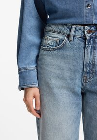 Jeans de mezclilla azul claro con cintura alta, textura desgastada y un bolsillo delantero que muestra un logo. Combinados con una camisa de mezclilla de manga larga.