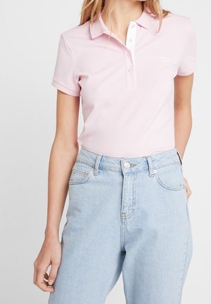 Vrouw die een lichtroze poloshirt met korte mouwen draagt, ingestopt in een hoog uitgesneden lichtblauwe spijkerbroek, staat met één hand in haar zak.