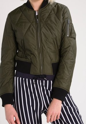 Person wearing a quilted olive green bomber jacket with black cuffs and waistband, paired with black and white vertical striped pants.