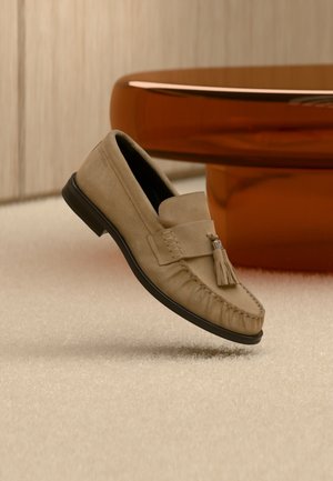 Mocasín de ante marrón claro con detalle de borla, suela negra, colocado sobre alfombra beige cerca de una base de mesa redonda marrón y brillante.