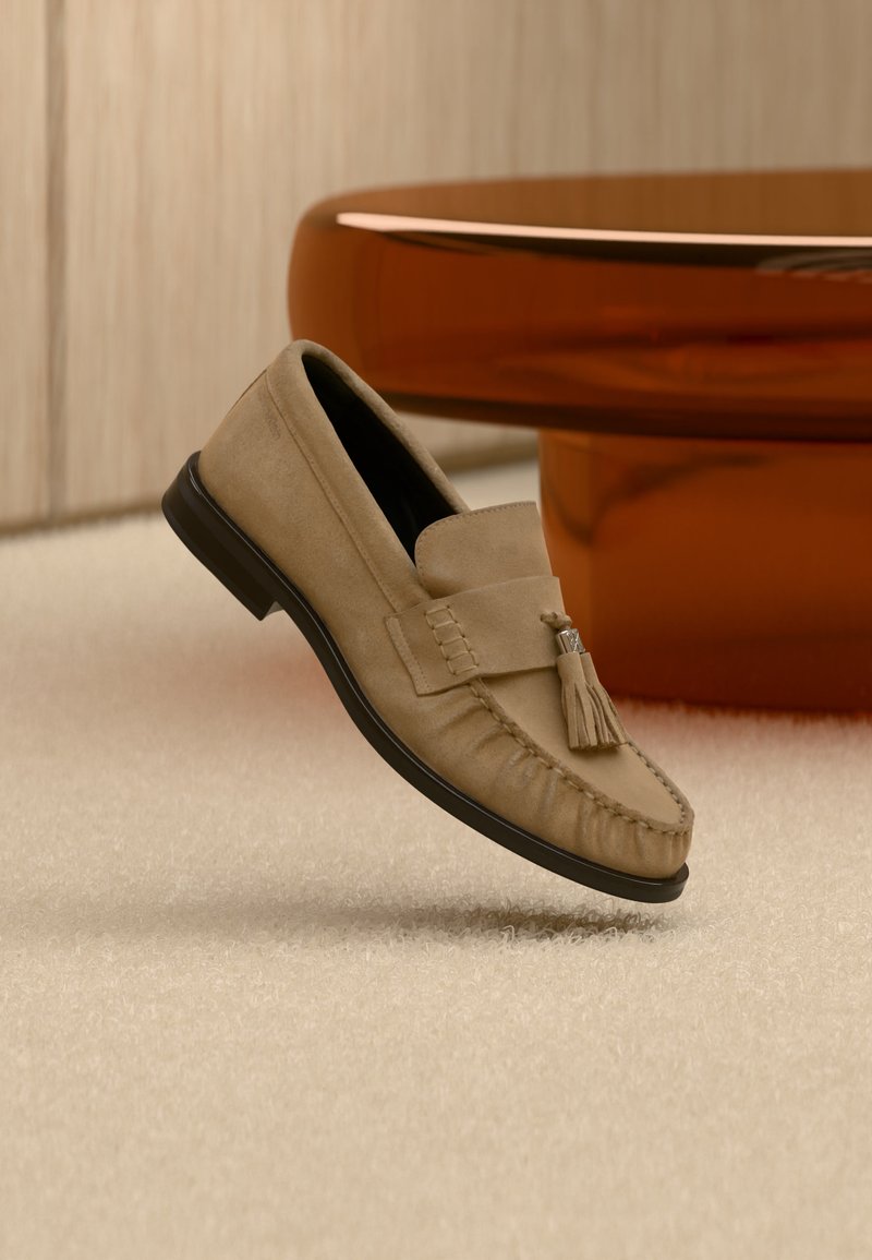 Ταμπά μοκασίνι από σουέτ με λεπτομέρεια φουντών, μαύρη σόλα, τοποθετημένο πάνω σε μπεζ χαλί κοντά σε γυαλιστερή καφέ στρογγυλή βάση τραπεζιού.